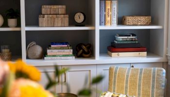 A cozy bookshelf display with books, plants, and baskets beside an armchair and brass lamp.