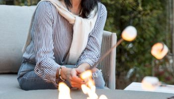 Smiling woman in a wide-brim hat relaxes on a porch beside a crackling fire pit in an outdoor garden.