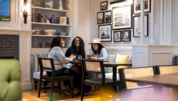 Three friends chat over drinks in a cozy, stylish dining room with bookshelves and framed photos on the walls.
