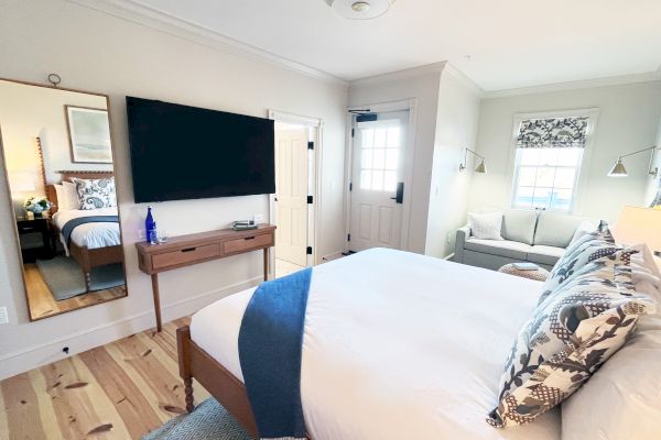 A modern bedroom with a bed, TV, mirror, and a small sofa. It has wooden floors, light walls, and a window bringing in natural light.