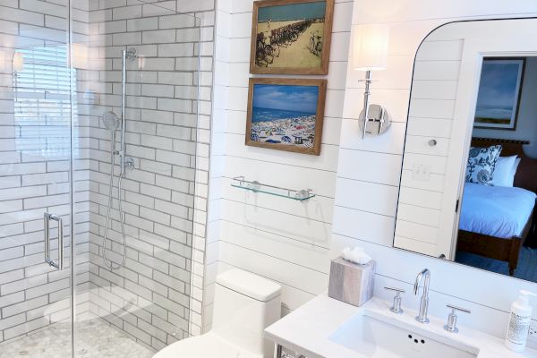 A modern bathroom with a glass shower, white tiles, a sink with a mirror, wall art, and a glimpse into a bedroom.