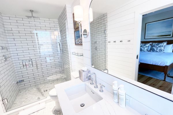 A bright bathroom with a glass shower, white subway tiles, a large mirror, and a view into a bedroom.