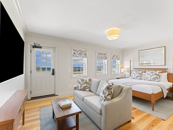 A cozy bedroom with a large bed, a sofa, a TV, and a small table. Three windows offer natural light, enhancing the airy atmosphere.