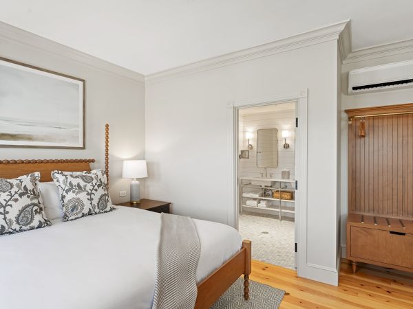 A cozy bedroom with a wooden bed, patterned pillows, a lamp, artwork, an open door to a bathroom, and a wooden wardrobe on the right.