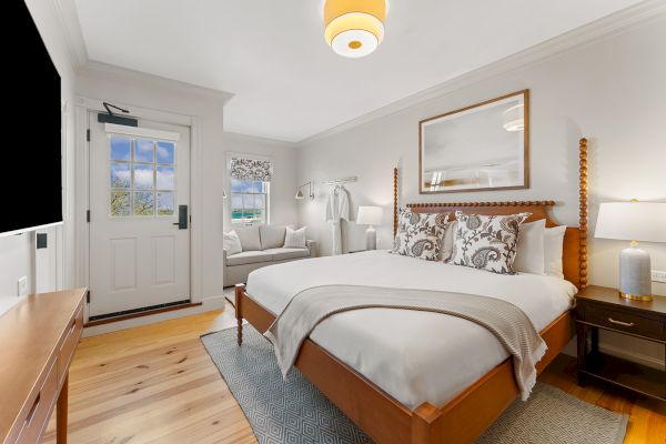 A cozy bedroom with a large bed, wooden furniture, a sofa, a framed picture, and a soft rug. Light streams through a window by the door.