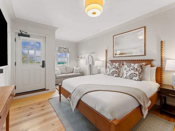 A cozy bedroom with a large bed, wooden furniture, a sofa, a framed picture, and a soft rug. Light streams through a window by the door.