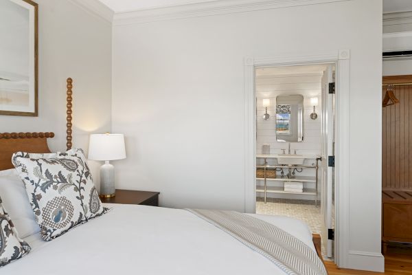 A cozy bedroom with a neatly made bed, decorative pillows, and a bedside lamp, leading to a bright bathroom with a mirror and double sinks.