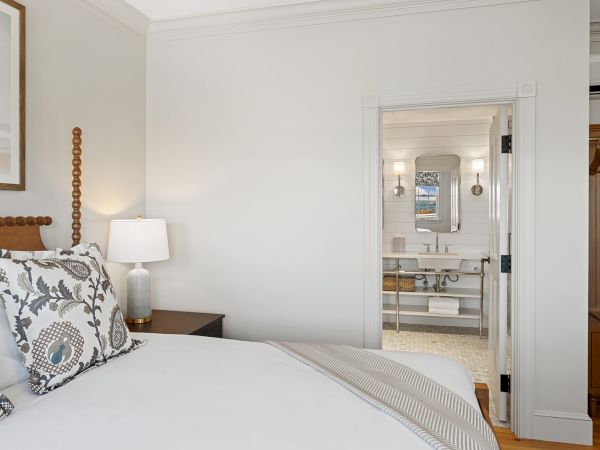 A cozy bedroom with a neatly made bed, decorative pillows, and a bedside lamp, leading to a bright bathroom with a mirror and double sinks.