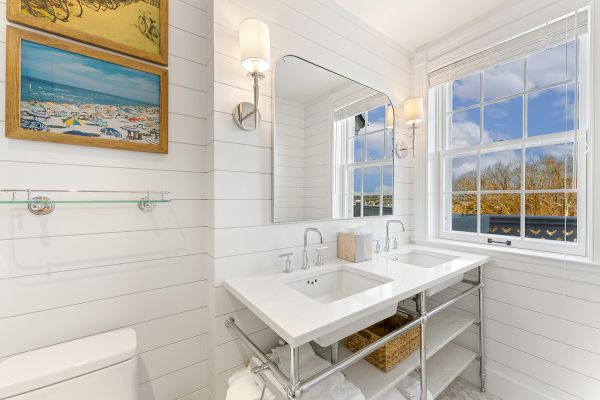 The image shows a bright bathroom with dual sinks, a large mirror, and a window. Wall art and light fixtures add decoration.