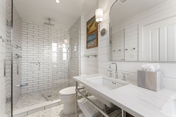 A modern bathroom features a glass shower, white tiles, a white vanity with a sink, a toilet, and minimalist decor, including wall art.