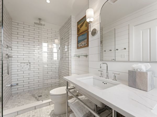 A modern bathroom features a glass shower, white tiles, a white vanity with a sink, a toilet, and minimalist decor, including wall art.