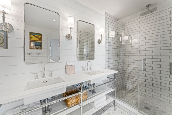 A modern bathroom with twin sinks, two mirrors, wall lamps, and a glass-enclosed shower. White tiles and wooden accents complete the decor.