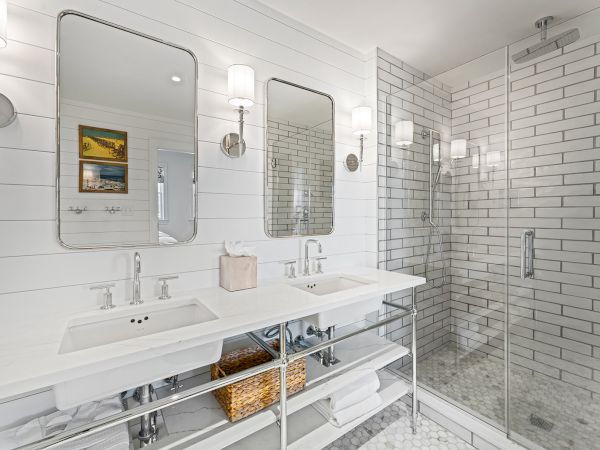 A modern bathroom with twin sinks, two mirrors, wall lamps, and a glass-enclosed shower. White tiles and wooden accents complete the decor.