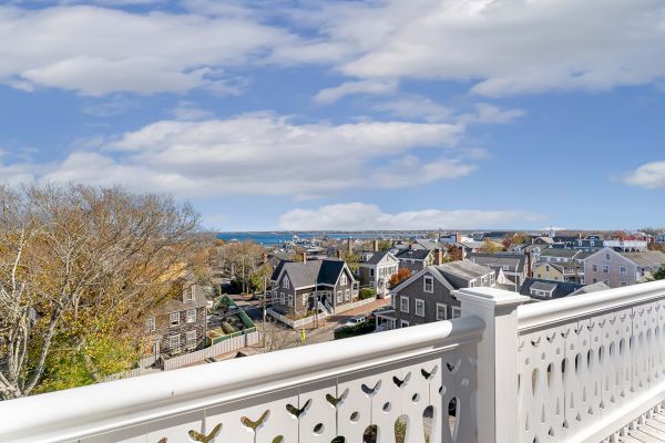 A scenic view of a coastal town with houses, trees, and a glimpse of the ocean, seen from a balcony with a white railing.