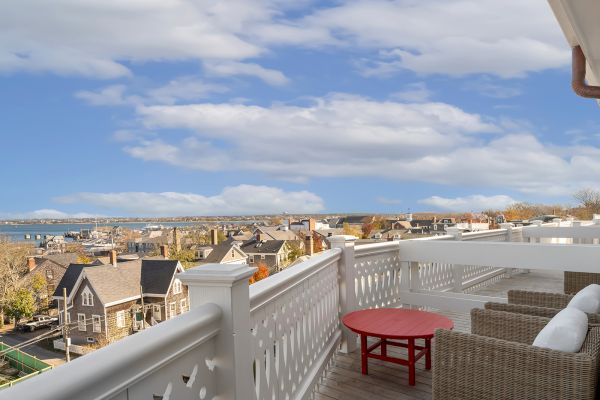 A scenic view from a balcony featuring a red table and wicker furniture, overlooking houses and a coastal landscape with a cloudy sky.