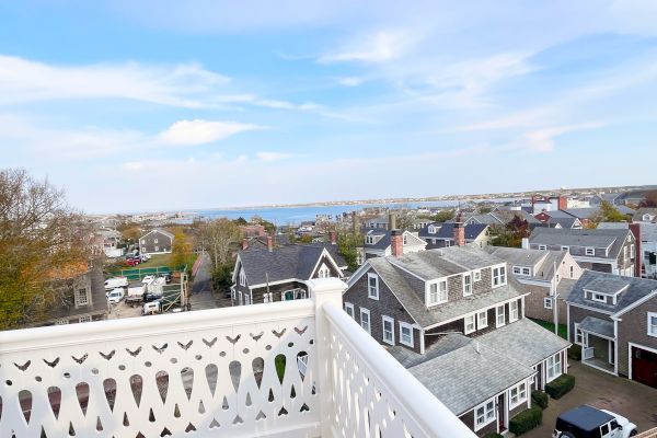 A view from a balcony shows a coastal town with classic houses and a glimpse of the sea under a clear blue sky.