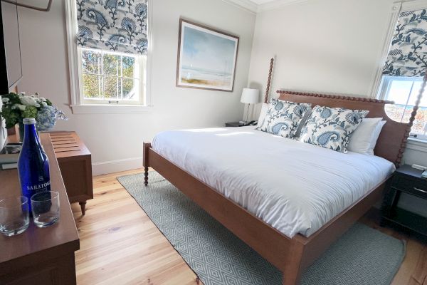 A cozy bedroom with a wooden bed, patterned pillows, floral blinds, a framed picture, and a wooden dresser with water bottles on top.