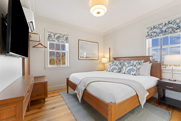 A cozy bedroom with a large bed, patterned pillows, a television, and windows with curtains. Warm lighting and wooden accents complement the decor.