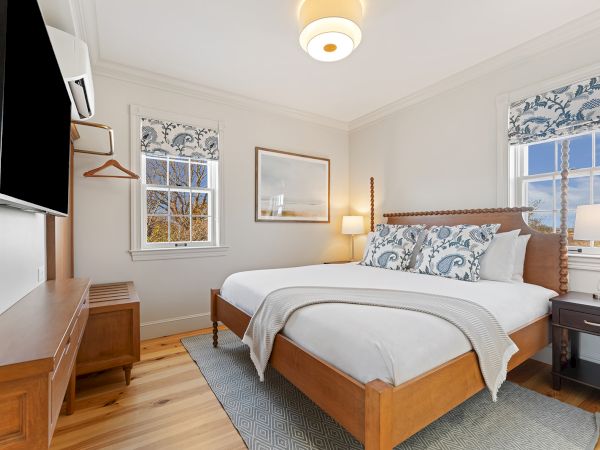 A cozy bedroom with a large bed, patterned pillows, a television, and windows with curtains. Warm lighting and wooden accents complement the decor.