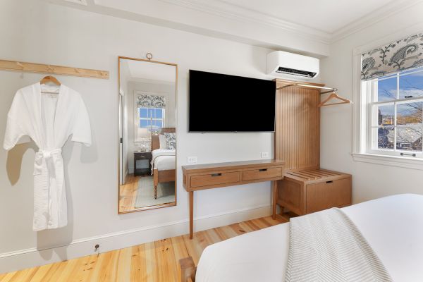 A bedroom features a wall-mounted TV, wooden dresser, mirror, clothing rack with a robe, and a window with patterned curtains.