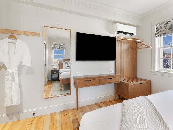 A bedroom features a wall-mounted TV, wooden dresser, mirror, clothing rack with a robe, and a window with patterned curtains.