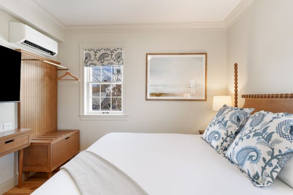 A cozy bedroom with a bed, blue-patterned pillows, wall art, and window. There's a wooden wardrobe, desk, and a mounted TV.