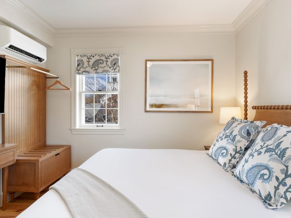 A cozy bedroom with a bed, blue-patterned pillows, wall art, and window. There's a wooden wardrobe, desk, and a mounted TV.