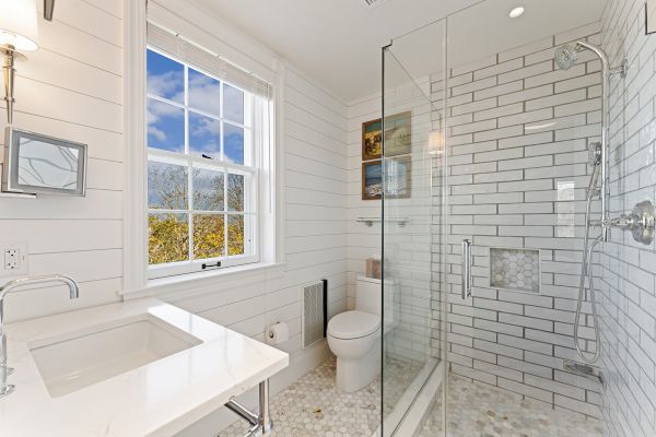 A bright, modern bathroom with a glass shower, white countertop, toilet, and a window with a view. White subway tiles cover the walls.