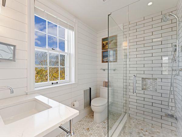 A bright, modern bathroom with a glass shower, white countertop, toilet, and a window with a view. White subway tiles cover the walls.