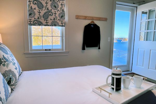 A cozy bedroom with a bed, a tray holding a coffee set, a window, a sweater on the wall, and an open door leading to a balcony with a view.