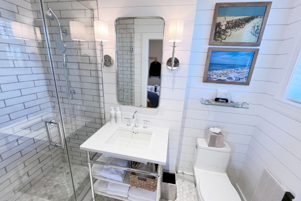 A modern bathroom with a glass shower, sink, wall mirror, two framed pictures, and a toilet on a white-tiled wall background.