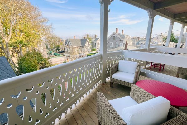 A balcony with wicker furniture overlooks a residential area with charming houses and trees under a blue sky.