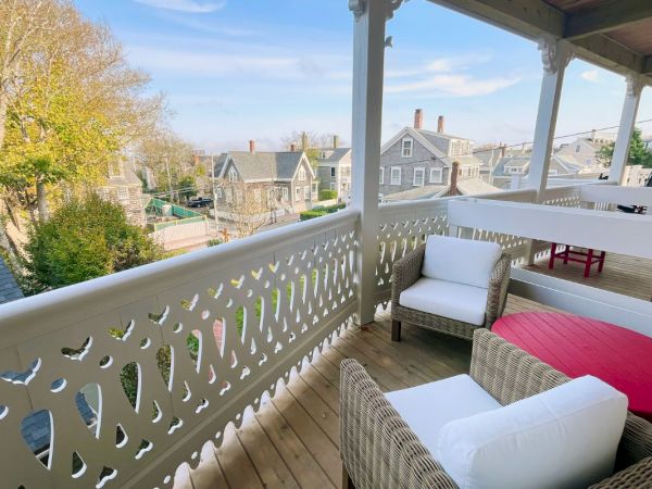 A cozy front porch with wicker seating, pillows, a red table, and a decorative white railing overlooking a neighborhood street.