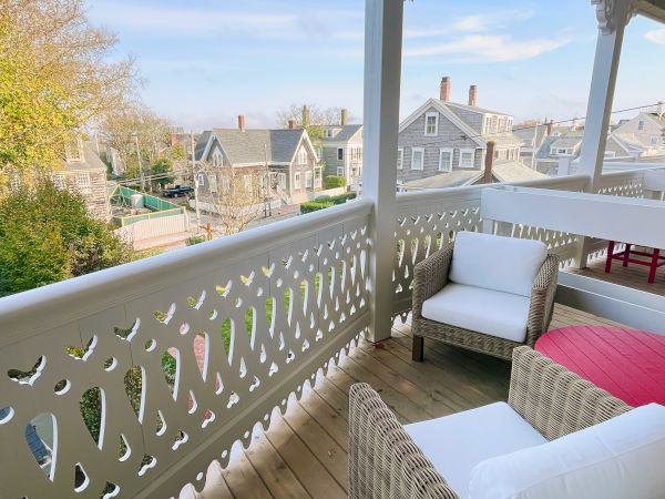 A porch with wicker furniture overlooks a charming neighborhood with classic houses, under a clear blue sky.