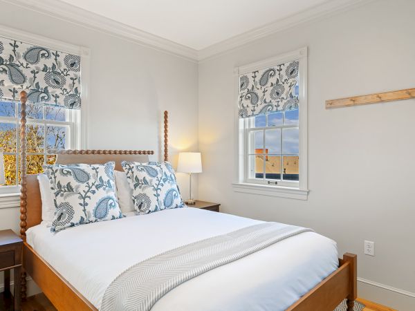 A cozy bedroom with a wooden bed, blue and white pillows, floral curtains, two windows, and a bedside lamp on a wooden nightstand.
