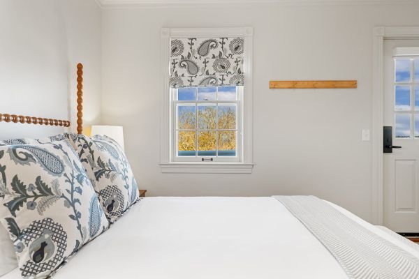 The image shows a bright bedroom with a bed, decorative pillows, a window with patterned curtains, and light entering the room.