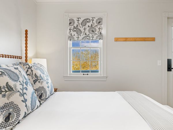 The image shows a bright bedroom with a bed, decorative pillows, a window with patterned curtains, and light entering the room.