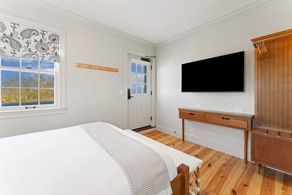 A bright bedroom with a bed, wall-mounted TV, desk, and window. The room has light wood flooring and a door with glass panels.