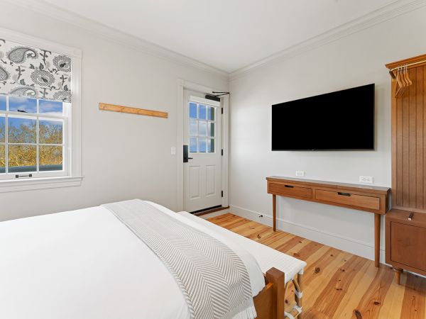A bright bedroom with a bed, wall-mounted TV, desk, and window. The room has light wood flooring and a door with glass panels.