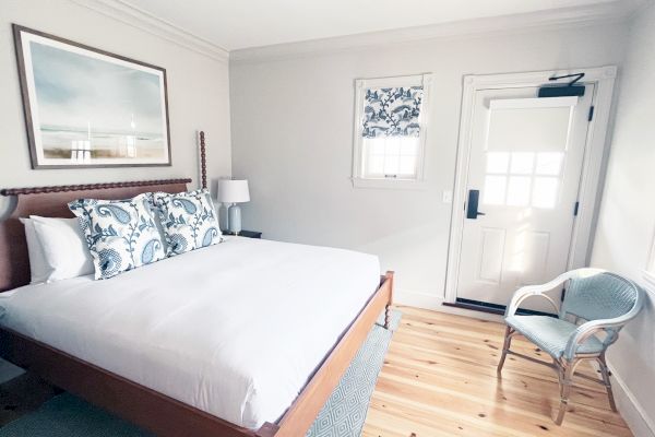 A minimalist bedroom with a large bed, decorative pillows, wooden floor, a chair, and artwork on the wall. There's a door and a small window.