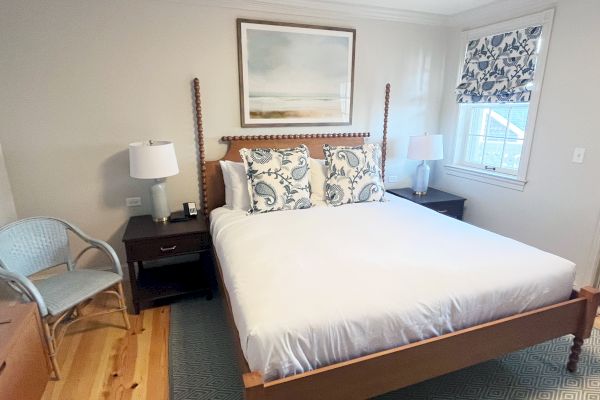 A cozy bedroom with a wooden bed, patterned pillows, nightstands with lamps, a chair, and a window with floral curtains. Peaceful decor.