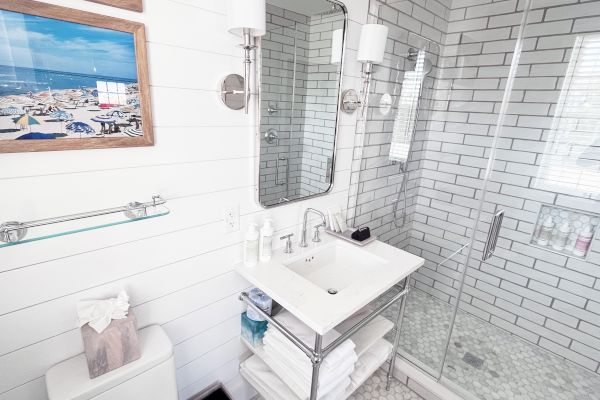 A modern bathroom with a glass shower, white tiles, a sink with towels below, a mirror, wall lights, and a coastal-themed picture.