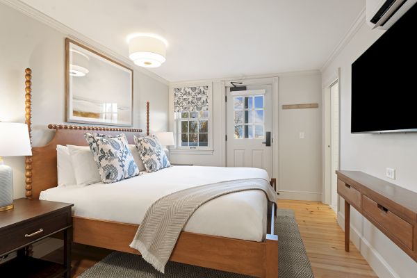 A cozy bedroom with a large bed, patterned pillows, and wall art. A TV is mounted on the wall, with a window and door nearby.