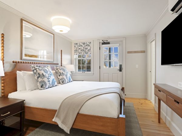 A cozy bedroom with a large bed, patterned pillows, and wall art. A TV is mounted on the wall, with a window and door nearby.