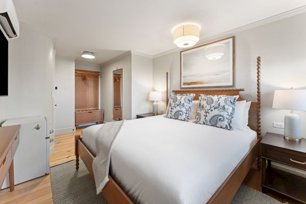 A cozy bedroom with a large bed, stylish pillows, modern lamps, and a painting above the headboard. Warm tones and soft lighting enhance it.