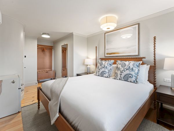 A cozy bedroom with a large bed, stylish pillows, modern lamps, and a painting above the headboard. Warm tones and soft lighting enhance it.