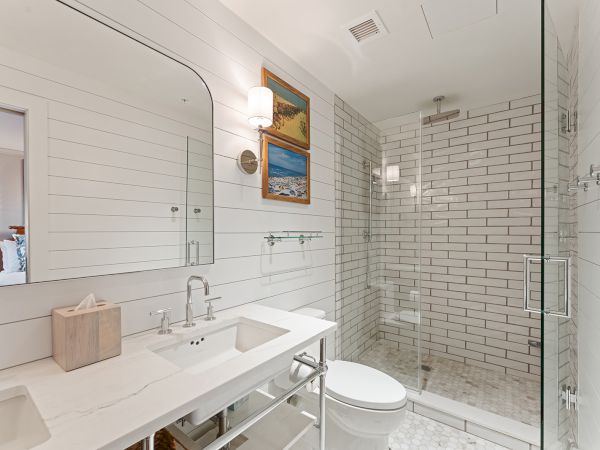 The image shows a modern bathroom with a white double sink vanity, a glass-enclosed shower with white tiles, and wall art above the toilet.