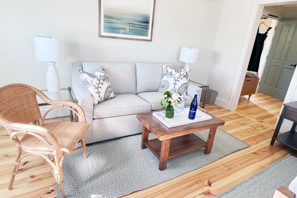 A cozy living room with a gray sofa, wicker chair, wooden table, decorative lamps, and a modern painting on the wall.