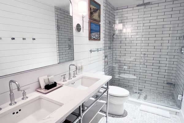 The image shows a modern bathroom with a double sink, large mirror, glass shower, and white subway tiles on the walls.