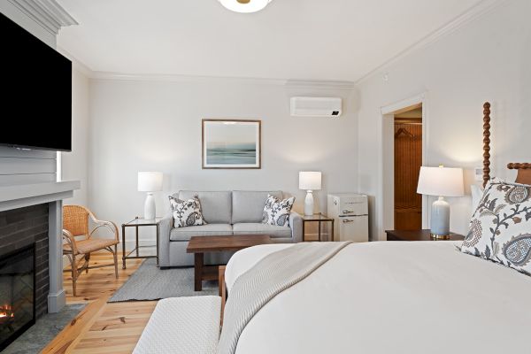 The image shows a cozy bedroom with a bed, sofa, TV, lamps, a fireplace, and a modern decor theme, featuring neutral tones and wooden accents.